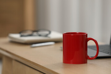 Red ceramic mug and laptop on wooden table at workplace. Space for text