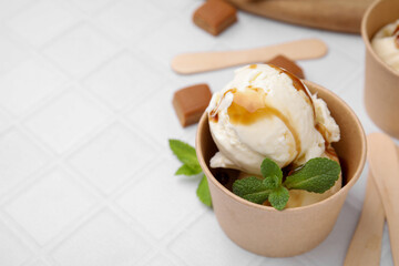 Scoops of ice cream with caramel sauce, mint leaves and candies on white tiled table, closeup. Space for text