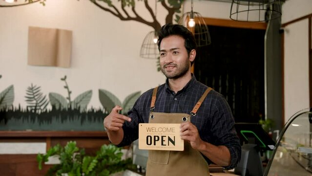 Indian man smiling wearing overalls standing in the middle of a coffee shop that is a small family business in the hand holding a sign that says open to let you know that the shop is open