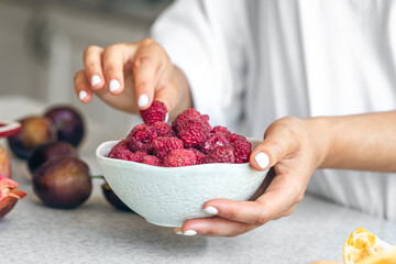 A bowl with raspberries in female hands on a white kitchen table.