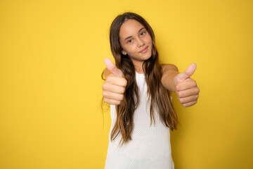 Little girl wearing a white dress showing thumb up on a yellow background.