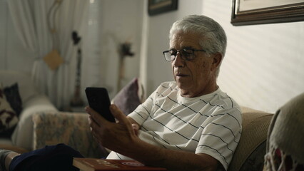 Elderly man staring at cellphone device sitting at home couch with concerned expression, watching video media online with baffled emotion