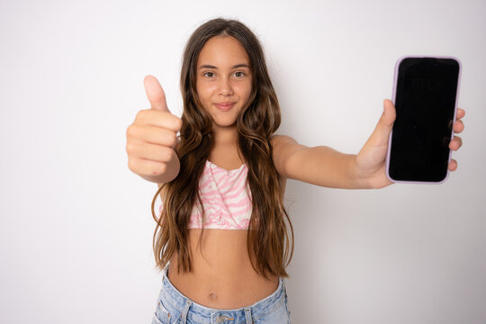 Smiling Little Kid Girl 11-12 Years Old In Summer Clothing Isolated On White Background . Childhood Lifestyle Concept. Mock Up Copy Space. Hold Mobile Phone With Blank Empty Screen, Showing Thumb Up