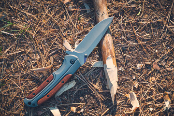 Folding hunting knife for survival and sharpened peg lie ground.