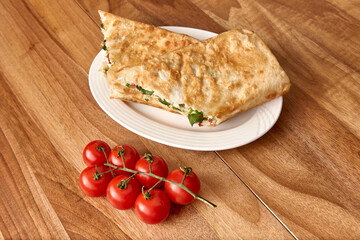 White plate with fried pita roll with salad with greens and tomatoes on the wooden table with cherry tomatoes near it