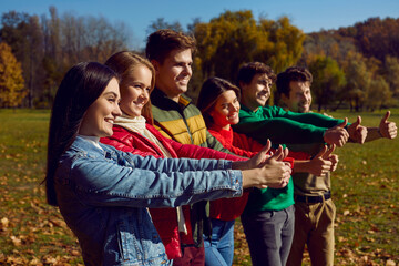 Group of happy people standing together showing thumbs up. Side view portrait of friends wearing casual clothes doing approval gesture in autumn park. Positive, optimistic, enthusiastic people