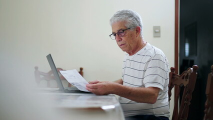 Upset elderly man in front of computer having to pay for vehicle fine sitting at home kitchen using laptop and typing on keyboard