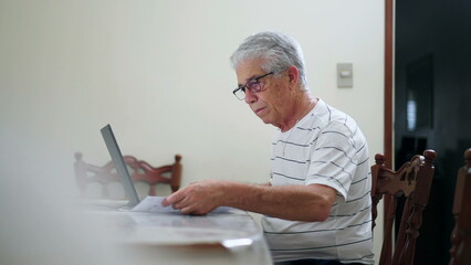 Upset elderly man in front of computer having to pay for vehicle fine sitting at home kitchen using laptop and typing on keyboard