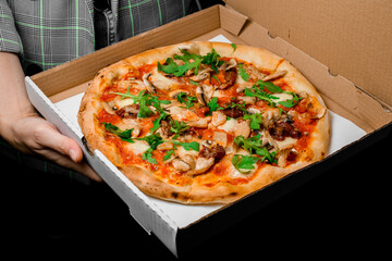 Restaurant manager holds packing box with fresh baked tasty Italian pizza with parmesan cheese, tomato sauce, sun dried tomatoes, chicken meat, champignons and rucola