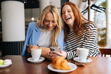 Portrait of two cheerful beautiful women friends sitting in cafe indoors, looking at phone and talking, laughing. Happy pretty females girlfriends using social media on smartphone in coffee shop.
