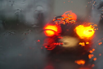 Abstraktes Motiv mit Wassermuster auf feuchter Glasscheibe vor Auto mit rot-gelben Lichtern auf Straße in Stadt bei Nebel, sturm und Regen am Morgen im Sommer