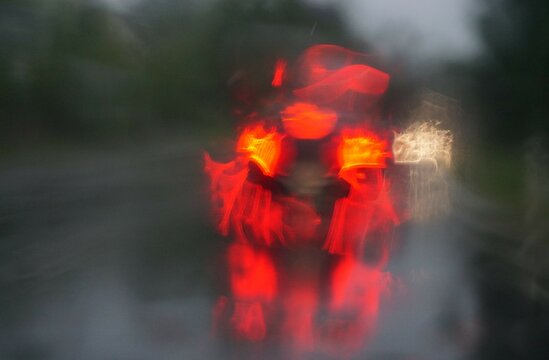 Abstraktes Motiv mit fahrendem Auto mit rot-gelben Lichtern auf nasser Straße vor Bäumen und Himmel bei Regen am Mittag im Sommer