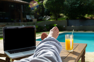 Mulher relaxando com seus pés em cima de uma mesa de madeira na beira da piscina com seu notebook ao lado e do outro um copo de suco de laranja, em dia de sol. © Madu Oliveira