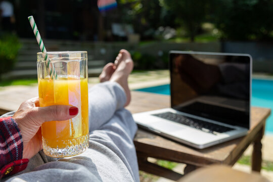 Mão de uma mulher segurando um copo de suco de laranja, vestida com calça de moletom cinza, com as pernas em cima da mesa e seu notebook ao lado, sentada perto da piscina.