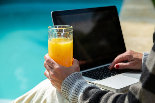 Mãos de uma mulher sentada à beira de uma piscina, trabalhando em seu notebook e segurando um copo de suco de laranja, em dia ensolarado.