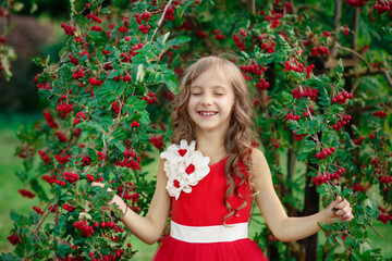 Young happy smiling girl in elegant red dress standing with closed eyes near tree with red berries of viburnum and laughing in summer park, emotional kid's portrait, idea of happiness and childhood 