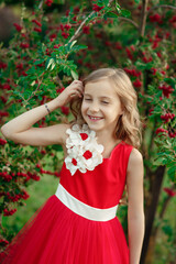 Obraz na płótnie Canvas Сute cheerful girl in red dress with flowers on it standing near tree with red berries, squinting and laughing in green park, vertical colorful outdoor summer kid's portrait, idea of happy childhood