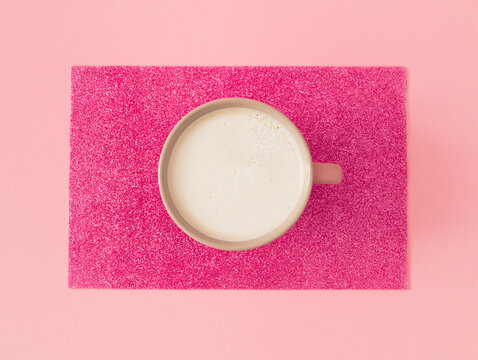 Think pink! Trendy arrangement of pink cup of coffee with coffee foam on pastel and sparkly pink background. Minimal coffee concept. Creative latte macchiato idea. Flat lay composition.