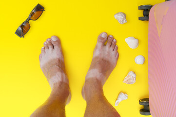 Tanned man's feet with seashells and suitcase on yellow background. Vacation card