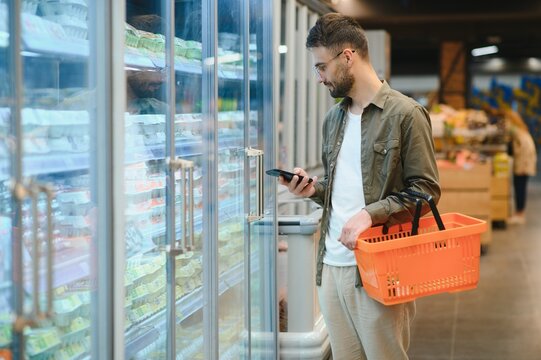 Quick Text During Shopping. Handsome Young Man Holding Mobile Phone And Smiling While Standing In A Food Store