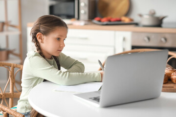 Little girl with laptop studying computer sciences online in kitchen