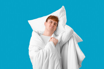 Young man with soft pillow and blanket on blue background