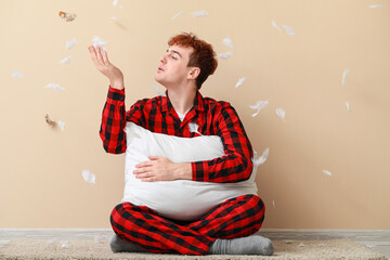 Young man with soft pillow and feathers sitting near beige wall