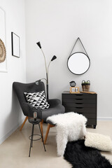 Interior of light living room with grey armchair, footstool, chest of drawers and mirror