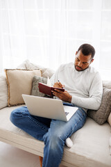 Young african american man writing on notebook near laptop while studying at home