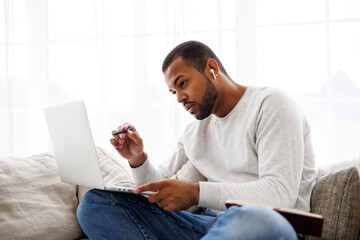 Focused african american student in earphone using laptop near blurred notebook on couch at home
