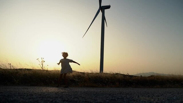 Silhouette of happy child girl spinning turn around hands to the sides on nature sunset in the field with windmill wind generator. Freedom, childhood, Environmentally friendly energy power.