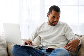 Young african american man in earphone looking at notebook while using laptop at home
