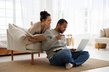 Young asian woman holding book near african american boyfriend pointing at laptop during online courses at home