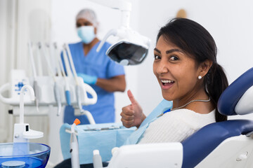 Smiling hispanic female patient showing thumb up after procedures in modern dental clinic