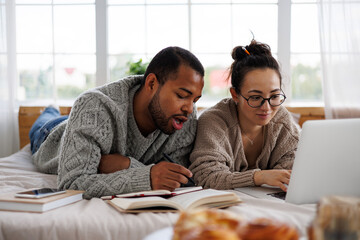 young multiracial couple in warm clothes using laptop and books during online courses on bed at home