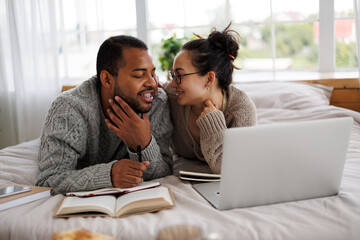 Smiling asian woman in eyeglasses hugging african american boyfriend during online education near laptop and books on bed 