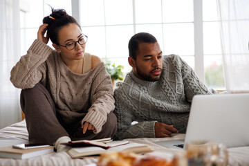 Young multiracial couple in sweaters using laptop and books during digital learning on bed at home