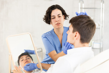 Obraz premium Young female cosmetologist consulting brunette guy lying in front of mirror at exam room of aesthetic center