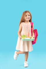 Happy little girl with backpack and lunchbox on blue background