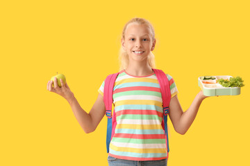 Happy girl with backpack, lunchbox and fresh apple on yellow background