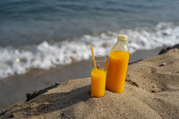 Fototapeta na wymiar a bottle and a glass of orange juice stand on the sand against the background of the sea