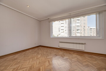 Empty living room with a large window with a view, a long aluminum radiator below, oak parquet floors and a white wooden door