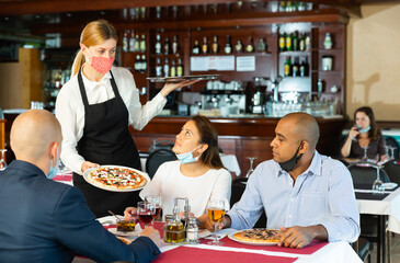 Waitress in protective mask serving delicious pizza to friends in cozy restaurant