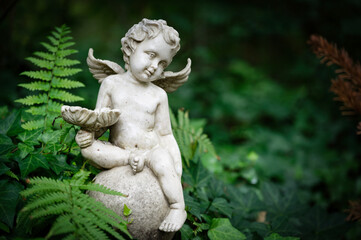 small putto with wings and bowl in hand sits on a ball on a grave surrounded by ferns