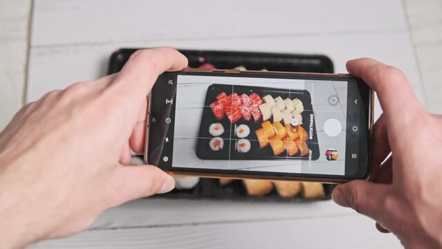 A male hands take a photo of sushi rolls in a delivery plastic box with a smartphone camera. Blogger shoots a review of Japanese food on a mobile phone for posting on social networks. Selective focus