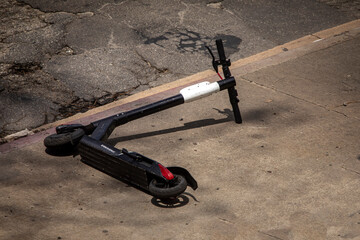 Public electric scooter laying on sidewalk near dirty poorly maintained street