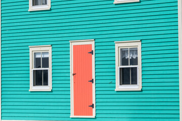 The exterior of a vibrant orange color single traditional wooden shutter door with cream color trim on a teal green clapboard wall of a historic house with two single hung windows with lace curtains.