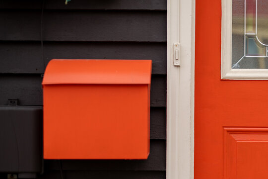 A square vibrant red metal mailbox on a navy blue wooden house with a vibrant red door. There's an electric doorbell between the postbox and the half-glass, half-metal door. 