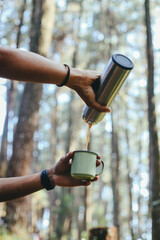 Human hand pours hot coffee or tea from a thermos into a mug on the mountain forest. Concept of autumn outdoor recreation. Low angle view