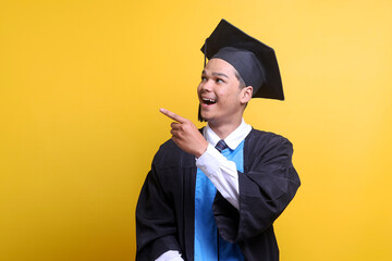 Excited Asian male in graduation clothes looking and pointing aside to the copy space for text or...